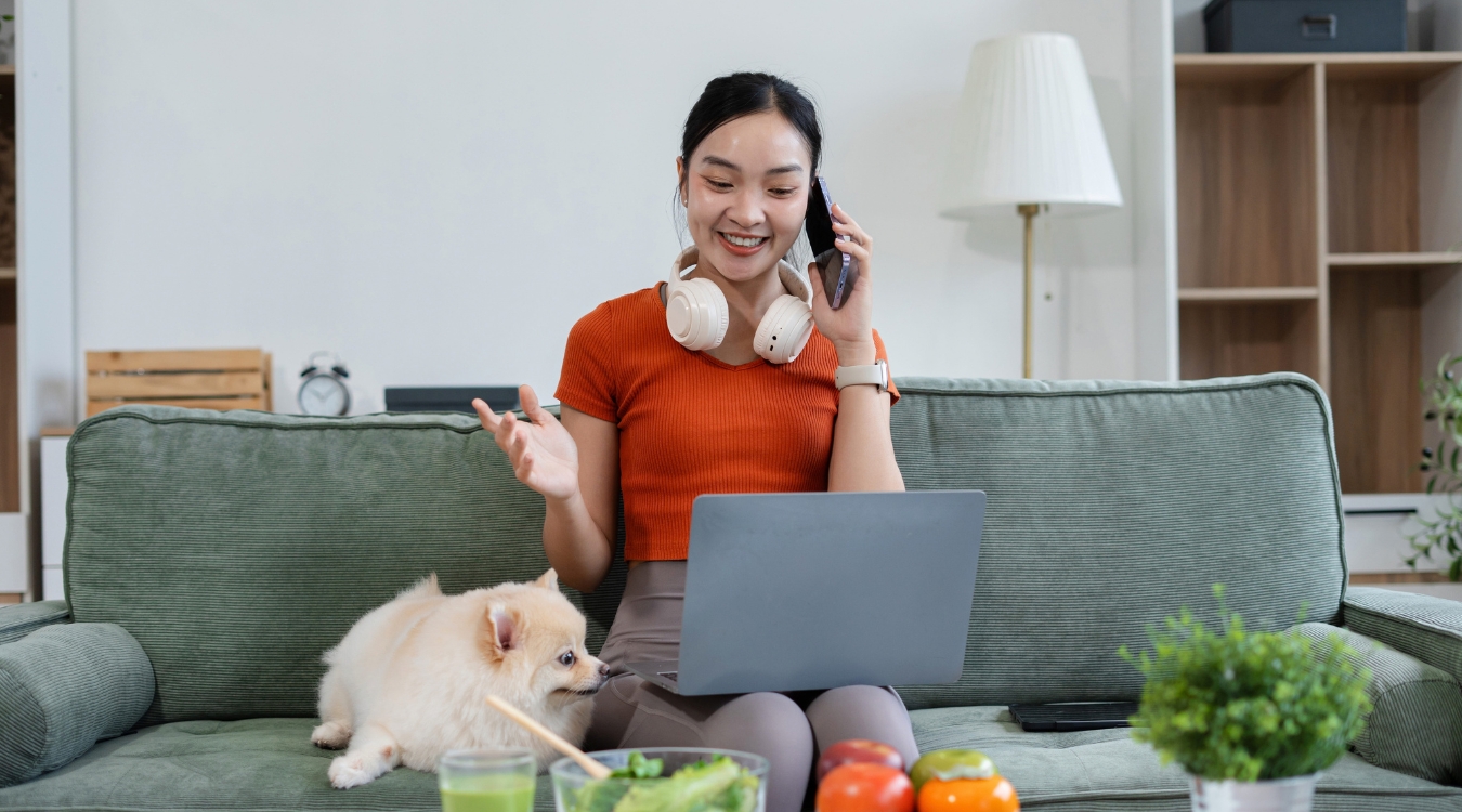 Filipina freelancer happily accepting freelance task on phone call while working on stable remote job on laptop sitting on sofa with dog beside her