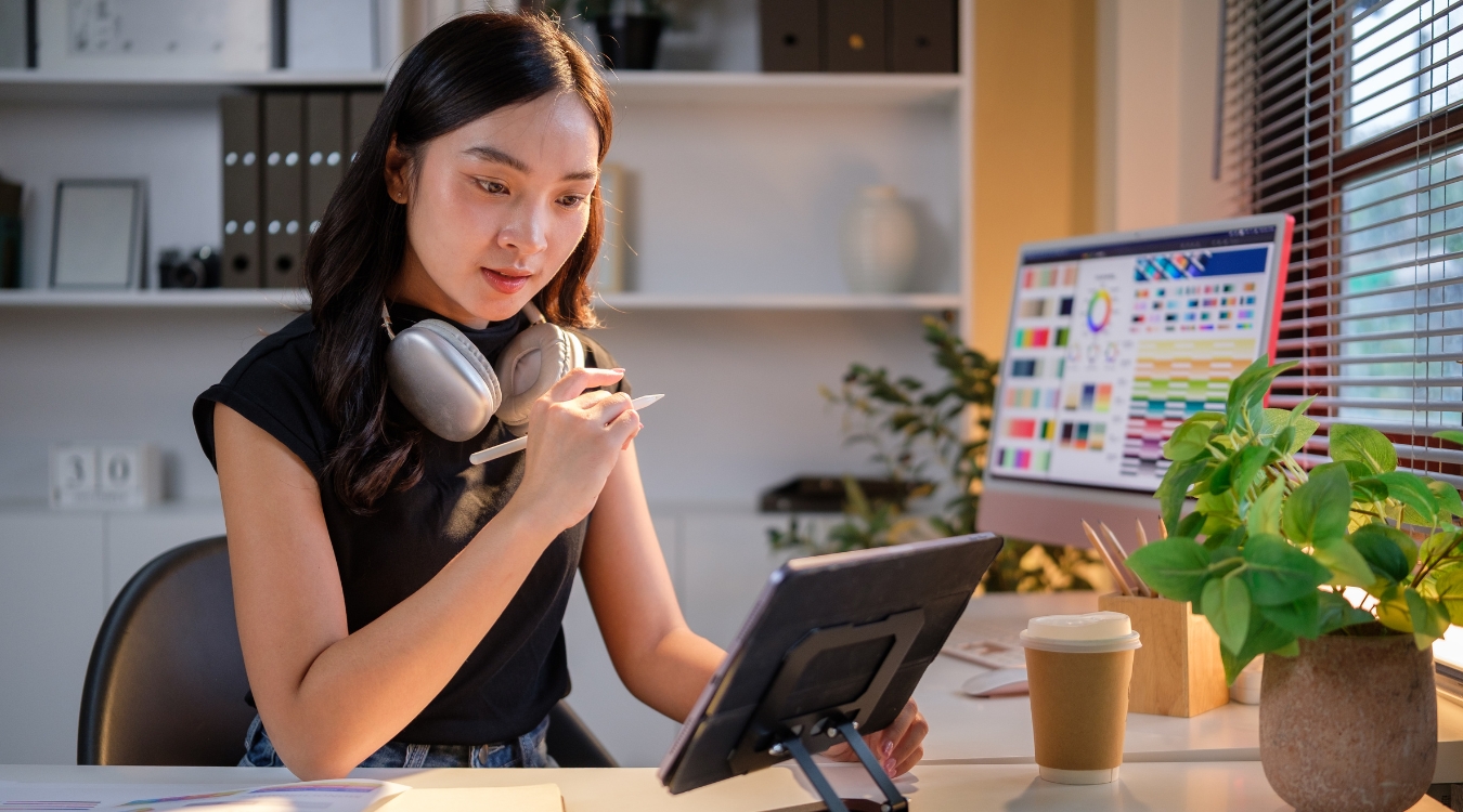 Filipina freelance graphic designer working on iPad with desktop computer showing color panels in home office setup